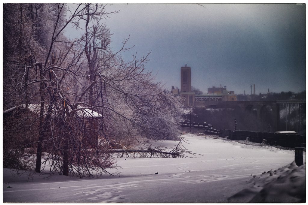 Snow-covered riverside path in Niagara Falls, Ontario, with frost-coated bare trees and a fallen branch in the foreground, and a misty bridge and buildings in the distance under a cold winter sky.