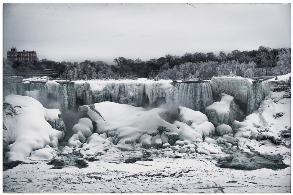 A wide winter view of the American Falls at Niagara Falls, seen from the Canadian side, with thick ice formations and snow-covered rocks surrounding the cascading water. Frozen mist clings to the cliff edges and trees along the rim, while a bridge and mid-rise building appear in the distance under an overcast sky.